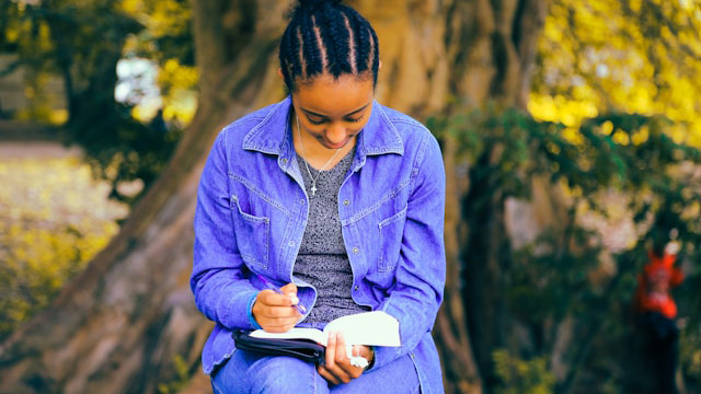 A young woman sitting on a park bench writing in a notebook